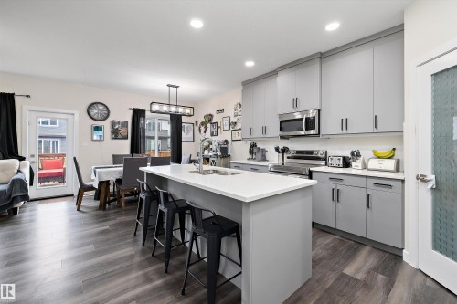 Open-concept kitchen and dining area featuring a central island with seating, grey cabinetry, stainless steel appliances, and wood-finish flooring - 84 Wingate Way, Fort Saskatchewan, AB 