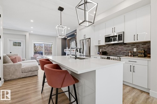 Modern kitchen featuring a large central island with seating, white shaker-style cabinetry, stainless steel appliances, and a mosaic tile backsplash - 61 230 Edgemont Road, Edmonton, AB - Indoor Photo Showing Kitchen With Double Sink With Upgraded Kitchen