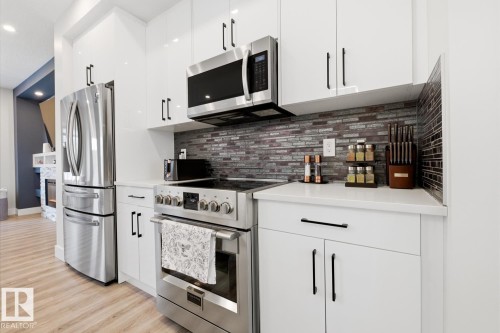 Contemporary kitchen featuring white cabinetry with black hardware, stainless steel appliances, white countertops, and a mosaic tile backsplash - 61 230 Edgemont Road, Edmonton, AB - Indoor Photo Showing Kitchen With Stainless Steel Kitchen With Upgraded Kitchen