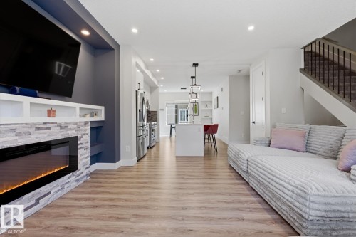 Open-concept living space featuring wood-finish flooring, a contemporary electric fireplace with stone-look surround, recessed lighting, a modern kitchen island with pendant lighting, and a staircase with dark balusters - 61 230 Edgemont Road, Edmonton, AB - Indoor Photo Showing Living Room With Fireplace