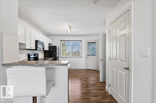 Kitchen featuring white cabinetry, black appliances, a breakfast bar, wood-finish flooring, and a window with blinds - 18 6032 38 Avenue, Edmonton, AB - Indoor Photo Showing Kitchen