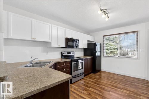 Kitchen area featuring wood-finish flooring, white cabinetry, dark lower cabinets, a dual basin stainless steel sink, and a window with blinds - 18 6032 38 Avenue, Edmonton, AB - Indoor Photo Showing Kitchen With Double Sink