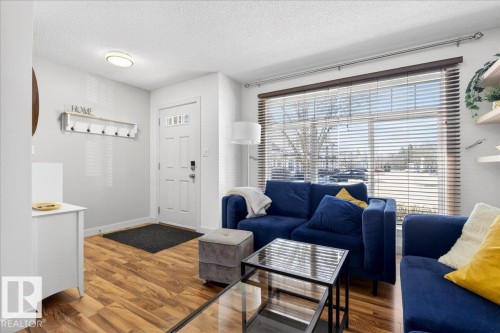 Entryway and living area featuring wood-finish flooring, a white paneled entry door with four glass inserts, and a large front window with horizontal blinds - 18 6032 38 Avenue, Edmonton, AB - Indoor