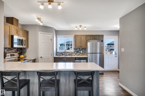 Kitchen featuring a peninsula with countertop seating, wood-finish cabinetry, stainless steel appliances, a tile backsplash, and a window above the sink - 2424 Wonnacott Crest, Edmonton, AB - Indoor Photo Showing Kitchen With Stainless Steel Kitchen With Upgraded Kitchen