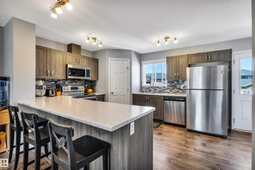 Kitchen featuring a large breakfast bar with light-toned countertop, wood-finish cabinetry, stainless steel appliances, a mosaic tile backsplash, and wood-finish flooring - 2424 Wonnacott Crest, Edmonton, AB - Indoor Photo Showing Kitchen With Stainless Steel Kitchen
