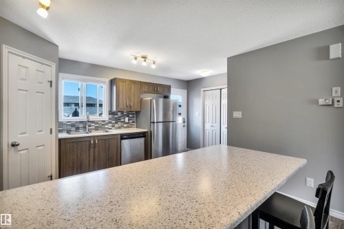 Kitchen featuring a spacious peninsula with a light speckled countertop, wood-finish cabinetry, a glass tile backsplash, a stainless steel refrigerator, and a stainless steel dishwasher - 2424 Wonnacott Crest, Edmonton, AB - Indoor Photo Showing Kitchen With Stainless Steel Kitchen