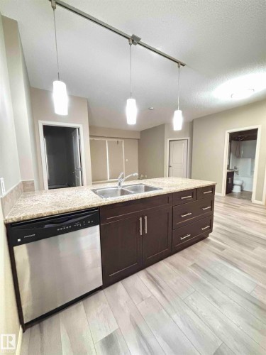 Kitchen island featuring a double basin stainless steel sink, granite countertop, dark wood cabinetry with chrome hardware, and an integrated stainless steel dishwasher - 408 111 Ambelside Drive Sw, Edmonton, AB - Indoor Photo Showing Kitchen With Double Sink