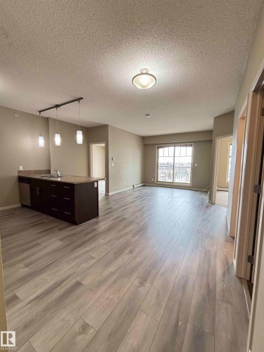 Open-concept living area featuring light wood-finish flooring, a kitchen island with dark cabinetry and a stone-finish countertop, a stainless steel dishwasher, and track pendant lighting - 408 111 Ambelside Drive Sw, Edmonton, AB - Indoor