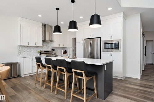 Contemporary kitchen featuring a spacious island with seating, white shaker cabinetry, stainless steel appliances, and wood-finish flooring - 591 Boulder Wynd, Leduc, AB - Indoor Photo Showing Kitchen With Upgraded Kitchen