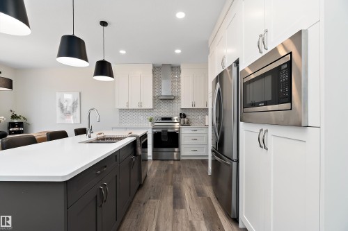 Contemporary kitchen featuring a large island with an undermount sink, white countertops, stainless steel appliances, white cabinetry, and wood-finish flooring - 591 Boulder Wynd, Leduc, AB - Indoor Photo Showing Kitchen With Double Sink With Upgraded Kitchen