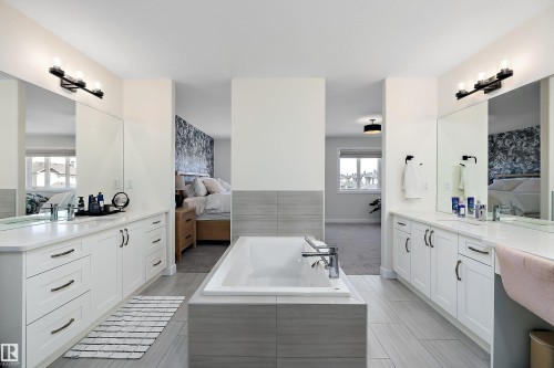 Freestanding soaking tub with a tiled surround, flanked by twin vanity stations featuring white cabinetry, integrated sinks, and individual mirrors with modern light fixtures - 591 Boulder Wynd, Leduc, AB - Indoor Photo Showing Bathroom