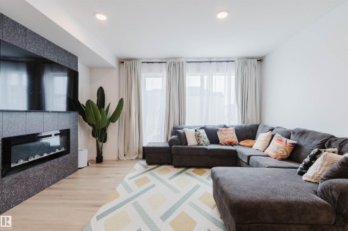 Living area featuring a textured accent wall with an integrated electric fireplace, light wood-finish flooring, recessed lighting, and multiple windows with sheer and opaque drapery - 3288 Keswick Way, Edmonton, AB - Indoor Photo Showing Living Room With Fireplace