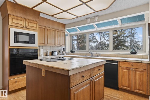Kitchen featuring wood-finish cabinetry, tile countertops, a built-in microwave and oven, a center island with an electric cooktop, and a window bank offering exterior views - 848 Wheeler Road W, Edmonton, AB - Indoor Photo Showing Kitchen