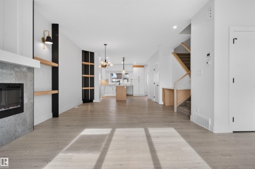 Open-concept living area featuring light wood-finish flooring, a built-in fireplace with a stone-finish surround, integrated shelving with brass sconces, a staircase with a wood-finish railing, and a kitchen with an island and pendant lighting - 45 River Birch, St. Albert, AB - Indoor With Fireplace