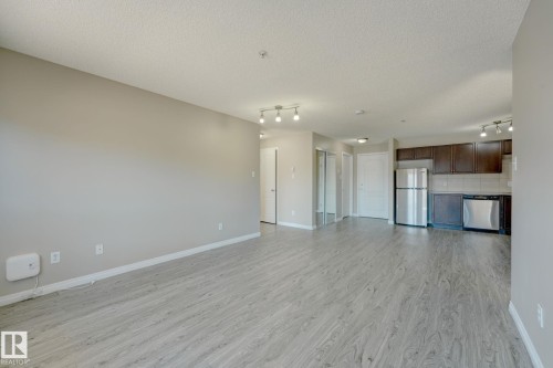 Spacious living area featuring light wood-finish flooring, neutral wall paint, and a kitchen with dark cabinetry and stainless steel appliances - 7226 7327 South Terwillegar Drive, Edmonton, AB - Indoor Photo Showing Other Room