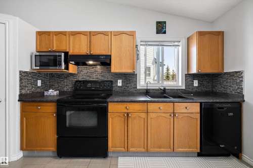 Kitchen featuring light wood cabinetry, dark countertops, a dark mosaic tile backsplash, black appliances, and a double-basin sink with a black faucet - 1811 37 Avenue, Edmonton, AB - Indoor Photo Showing Kitchen With Double Sink