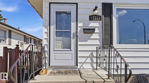 White siding exterior featuring a storm door with decorative glass, black wrought iron handrails, a contrasting black louvered shutter, an exterior wall light, and a prominent house number - 13533 119 Street, Edmonton, AB - Outdoor
