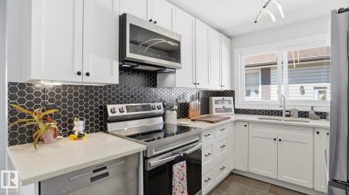 White cabinetry with dark hardware, a hexagonal tile backsplash, stainless steel appliances, light-toned countertops, and a window above the sink - 13533 119 Street, Edmonton, AB - Indoor Photo Showing Kitchen With Stainless Steel Kitchen