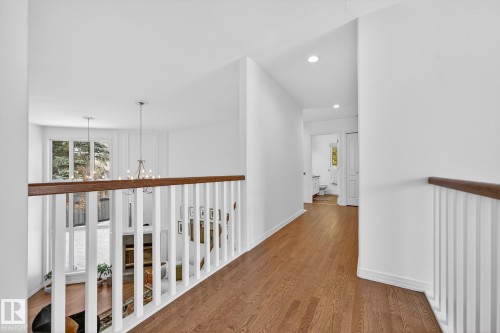Bright hallway with hardwood floors and white walls, featuring a wooden handrail and white balusters overlooking a lower level with large windows and a chandelier - 1211 Hewgill Place, Edmonton, AB - Indoor Photo Showing Other Room
