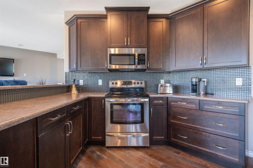 Kitchen featuring dark wood-finish cabinetry with crown molding, stainless steel appliances, a mosaic tile backsplash, and wood-finish flooring - 121 Ellington Crescent, St. Albert, AB - Indoor Photo Showing Kitchen