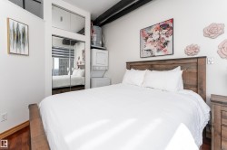 Bedroom featuring wood-finish flooring, a wood headboard, mirrored closet doors, upper mirrored storage, and a stacked washer/dryer unit - 