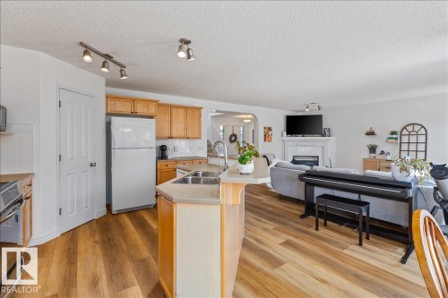 Open-concept living space featuring wood-finish flooring, a kitchen island with a double basin sink, white appliances, light wood cabinetry, and a fireplace with a mantel - 1130 Westerra Way, Stony Plain, AB - Indoor Photo Showing Kitchen With Double Sink