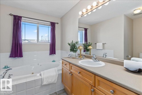 Bathroom featuring a built-in jetted tub with white tile surround, a window with grid detailing, and a vanity with wood-finish cabinetry, an integrated sink, and a large wall mirror with a Hollywood-style light fixture - 1130 Westerra Way, Stony Plain, AB - Indoor Photo Showing Bathroom