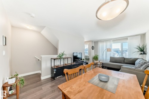 Open-concept living area featuring wood-finish flooring, a contemporary ceiling light fixture, a staircase with white railing, and bright windows with vertical blinds - 115 401 Southfork Drive, Leduc, AB - Indoor