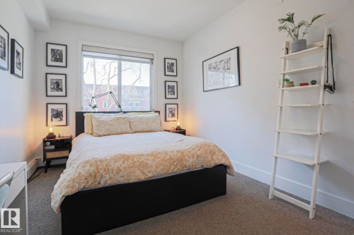 Bright bedroom featuring a large window with roller shades, neutral carpeting, and a built-in wall-mounted shelving unit - 202 10030 83 Avenue, Edmonton, AB - Indoor Photo Showing Bedroom