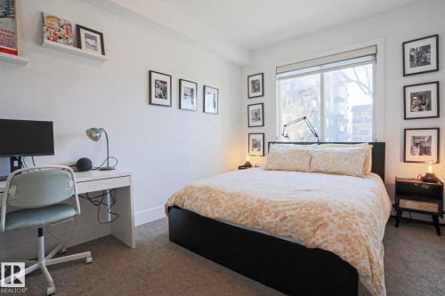 Bedroom featuring a large window with roller shade, neutral carpet flooring, white painted walls, and white baseboards - 202 10030 83 Avenue, Edmonton, AB - Indoor Photo Showing Bedroom