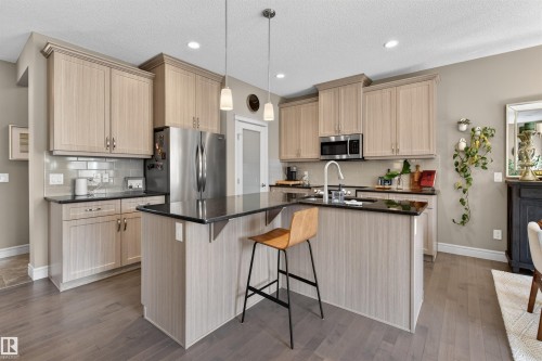 Modern kitchen featuring a central island with an integrated breakfast bar, light wood-finish cabinetry, stainless steel appliances, dark countertops, and a white subway tile backsplash - 2313 Sparrow Crescent, Edmonton, AB - Indoor Photo Showing Kitchen With Upgraded Kitchen