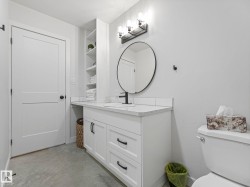 Bathroom featuring a white vanity with a white countertop, built-in shelving, a circular mirror with a black frame, and a modern three-light fixture - 