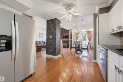 Open concept living space featuring wood-finish flooring, a kitchen with white cabinetry and dark countertops, a dining area with a modern light fixture, a brick-textured pillar, and a living area with a fireplace - 