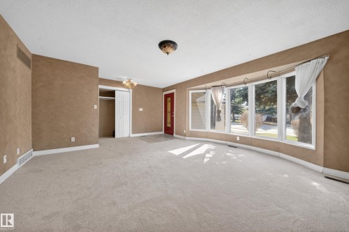 Spacious room featuring a curved bay window, light-toned carpet flooring, textured wall finishes, and a red entry door - 1767 61 Street, Edmonton, AB - Indoor Photo Showing Other Room