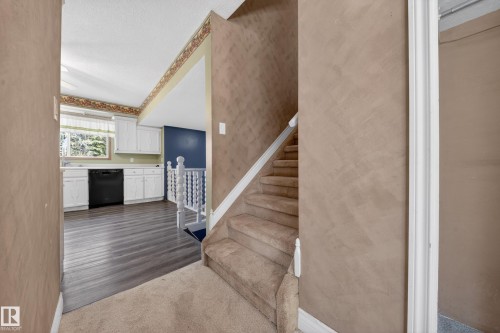 Carpeted staircase with white balusters leading to an open kitchen featuring wood-finish flooring, white cabinetry, and a black dishwasher - 1767 61 Street, Edmonton, AB - Indoor