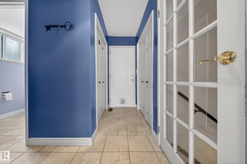 Interior hallway featuring square tile flooring, blue wall finishes, white baseboards, and white doors, including a French paneled door with a brass-finish handle - 1767 61 Street, Edmonton, AB - Indoor