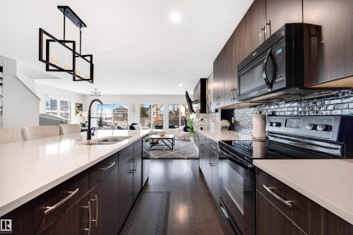 Contemporary kitchen with dark wood-finish cabinetry, light-toned countertops, integrated black appliances, a reflective tile backsplash, and a modern island pendant light - 2504 Coughlan Road, Edmonton, AB - Indoor Photo Showing Kitchen With Double Sink With Upgraded Kitchen