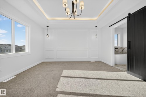 Spacious room featuring neutral carpeting, a large window, decorative wall paneling, a tray ceiling with recessed lighting and a central chandelier, and a black barn door opening to an adjacent space with a built-in tub and tiled surround - 3285 Chernowski Way, Edmonton, AB - Indoor Photo Showing Other Room