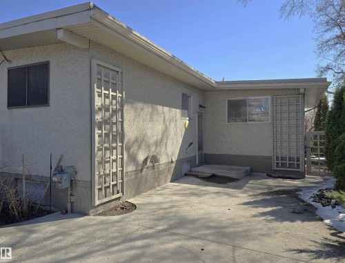Exterior of a stucco-finished property featuring a concrete patio, decorative lattice panels, and mature evergreens - 9624 75 Street, Edmonton, AB - Outdoor With Exterior