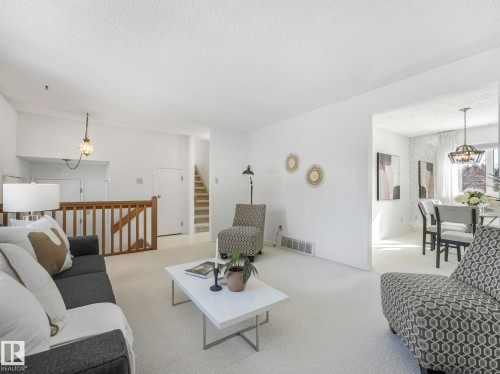 Spacious living area with light-colored carpeting and white walls, featuring a wooden railing and a view into a separate dining area with a chandelier - 3421 112A Street, Edmonton, AB - Indoor Photo Showing Living Room