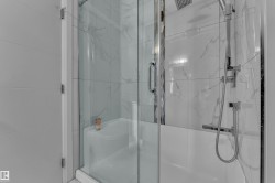 Enclosed shower and tub combination featuring clear glass doors, white tiled walls with subtle veining, a rainfall showerhead, and a handheld shower sprayer - 