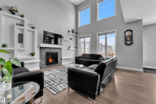 Vaulted ceiling living area featuring a stacked stone fireplace with a white mantel, custom wall shelving, multiple large windows, and wood-finish flooring - 3702 Claxton Place, Edmonton, AB - Indoor Photo Showing Living Room With Fireplace