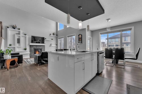 Open-concept living area featuring wide plank wood-finish flooring, a white kitchen island with an undermount sink, a stone-clad fireplace, and an accent wall with integrated shelving - 3702 Claxton Place, Edmonton, AB - Indoor Photo Showing Kitchen With Upgraded Kitchen