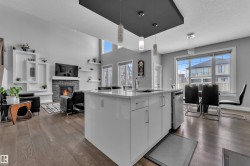 Open-concept living area featuring wide plank wood-finish flooring, a white kitchen island with an undermount sink, a stone-clad fireplace, and an accent wall with integrated shelving - 