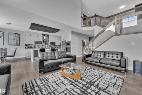 Open-concept living area featuring wood-finish flooring, a contemporary kitchen with white cabinetry and a mosaic tile backsplash, a kitchen island with seating, a dining area, and a glass-paneled staircase with wood accents - 3702 Claxton Place, Edmonton, AB - Indoor Photo Showing Living Room