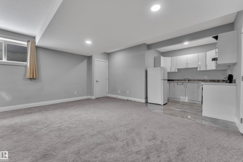 Open-concept living space featuring light gray carpet, recessed lighting, a compact kitchen with white cabinetry, a refrigerator, and a window with blinds - 3702 Claxton Place, Edmonton, AB - Indoor