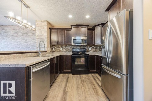 Kitchen featuring dark wood cabinetry, stainless steel appliances, stone-look countertops, a mosaic tile backsplash, and wood-finish flooring - 14 9350 211 Street, Edmonton, AB - Indoor Photo Showing Kitchen With Double Sink With Upgraded Kitchen
