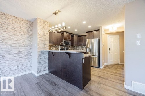 Open-concept kitchen featuring wood-finish flooring, dark wood cabinetry, stainless steel appliances, a breakfast bar, and a decorative pendant light fixture - 14 9350 211 Street, Edmonton, AB - Indoor Photo Showing Kitchen With Upgraded Kitchen