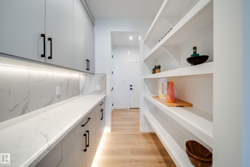 Modern pantry featuring extensive built-in shelving, white flat-panel cabinetry with black hardware, white stone-finish countertops, under-cabinet lighting, and light wood-finish flooring - 16 Blackbird Bend, Fort Saskatchewan, AB - Indoor