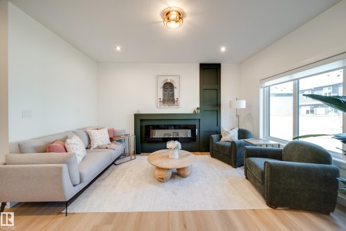 Spacious living area featuring light wood-finish flooring and a central fireplace with dark green surround - 16 Blackbird Bend, Fort Saskatchewan, AB - Indoor Photo Showing Living Room With Fireplace